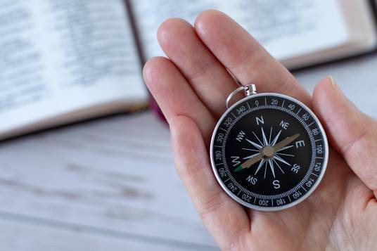Person hält Kompass mit einem aufgeschlagenen Buch unscharf im Hintergrund